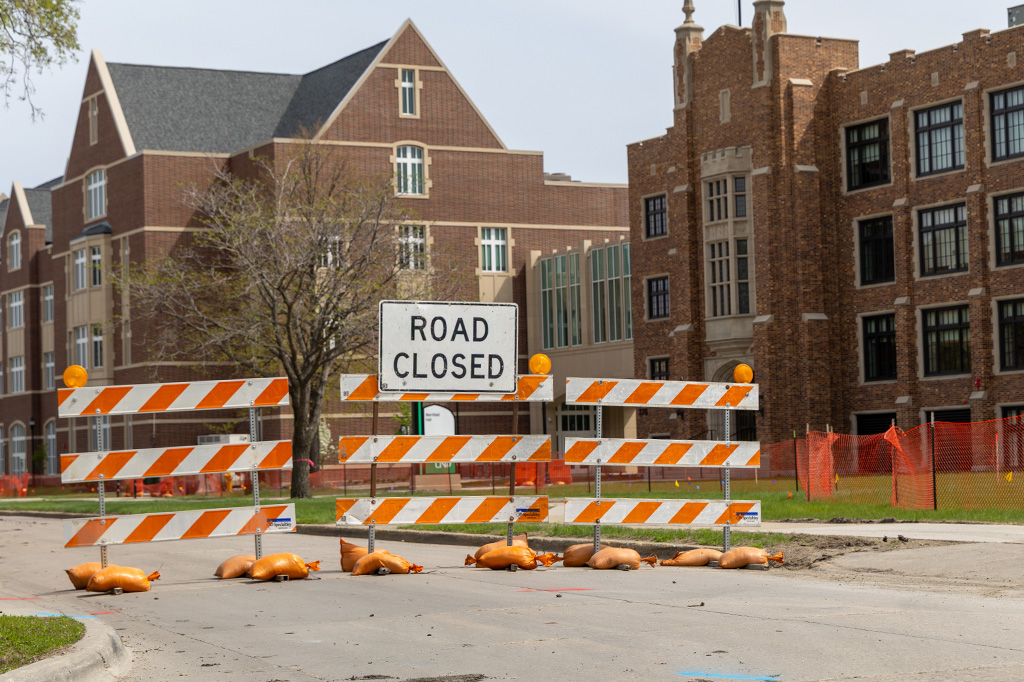 A sign reading "Road Closed" sits in the middle of a road with two campus buildings in background