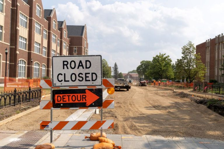 Traffic sign including road closed and detour arrow stand in front of road construction site, buildings to either side