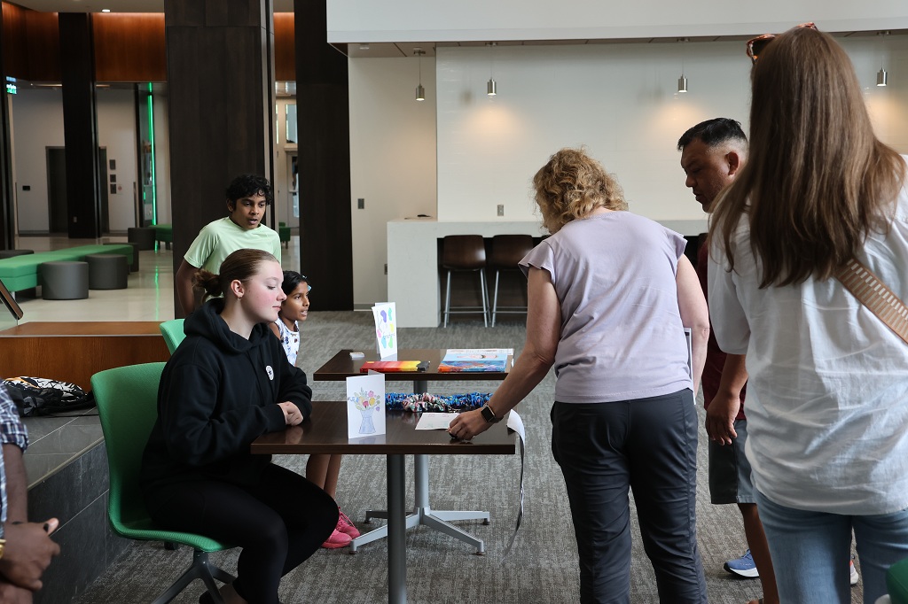 While sitting at a table that displays the products she has made, a young entrepreneur talks with a potential customer. 