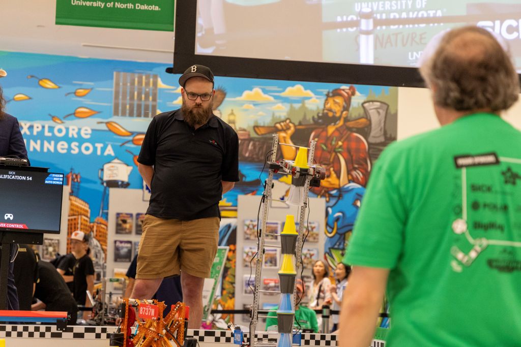 A VEX Robotics official watches as a robot lifts and stacks multicolored cones during a qualification match.