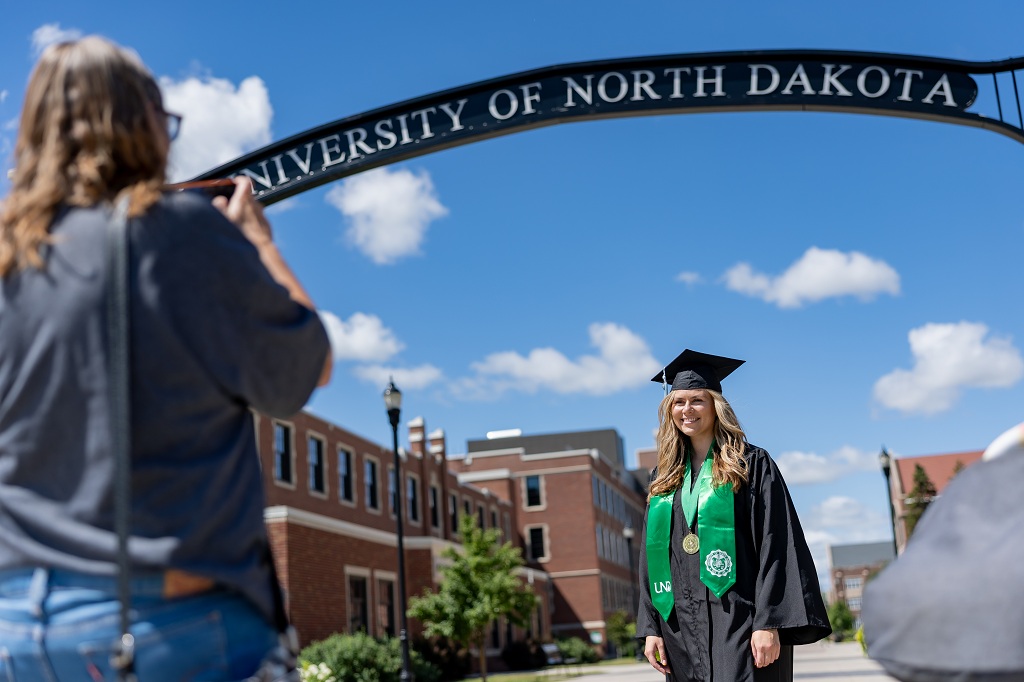Graduate wearing cap and gown poses for photo beneath UND arch