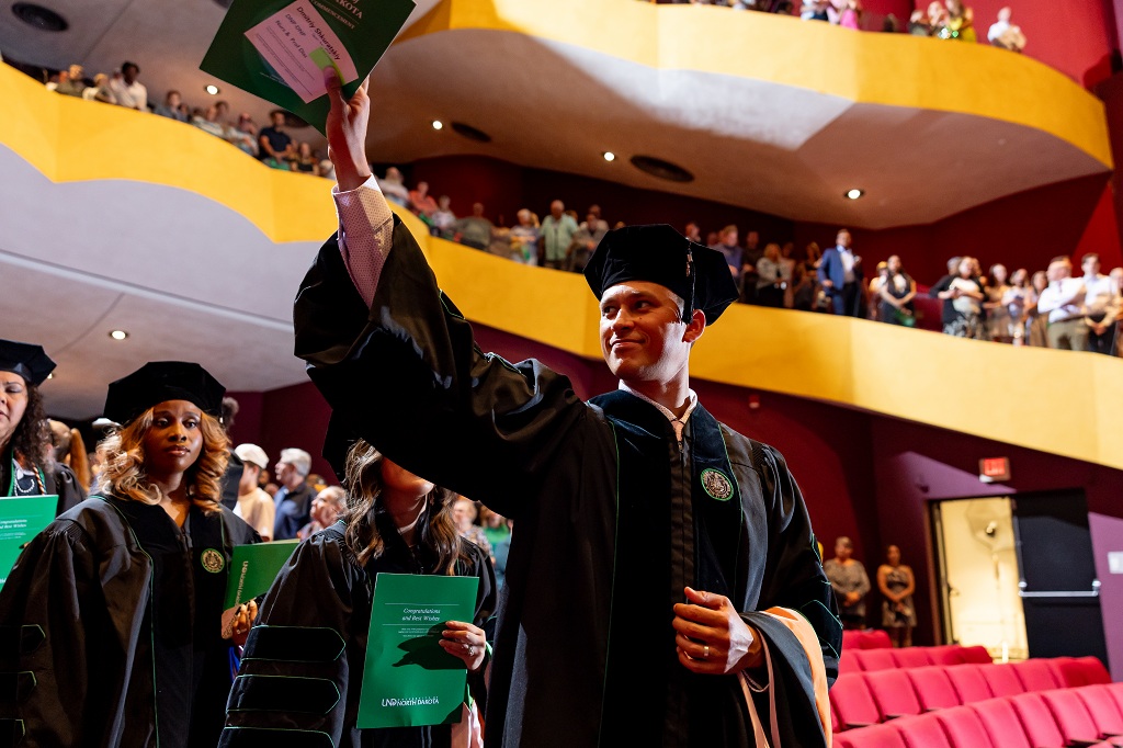 Man wearing cap and gown waves to Commencement crowd in auditorium