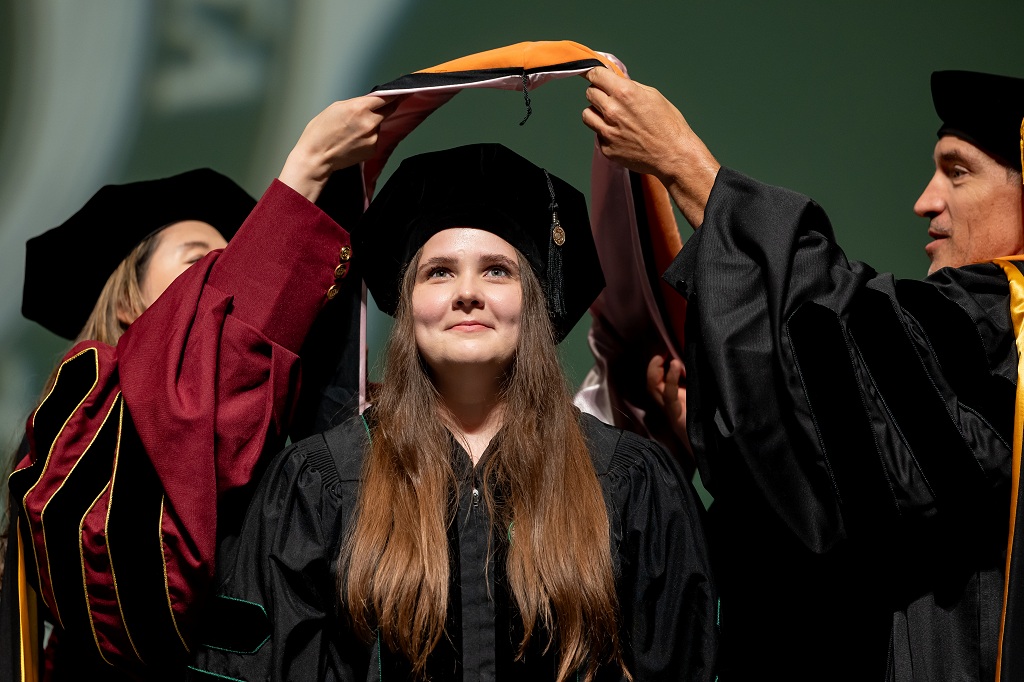 A woman wearing a cap and gown has a medallion hung around her neck at Summer Commencement 2026.