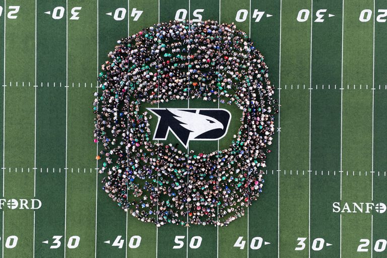 Aerial view of UND students gathered in a large circle around the Fighting Hawks logo at midfield inside the Alerus Center