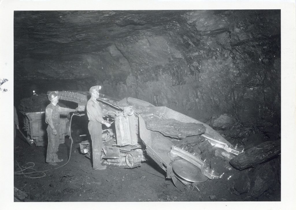Historic black-and-white photograph of two coal miners operating a conveyor that carries large chunks of lignite coal inside the Knife River Coal Mining Co. mine near Beulah, North Dakota.