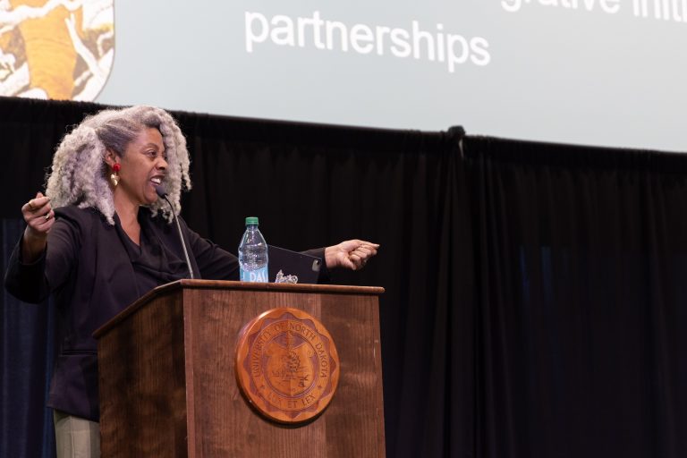 A woman speaks energetically from behind a UND podium, extending both arms toward the audience.