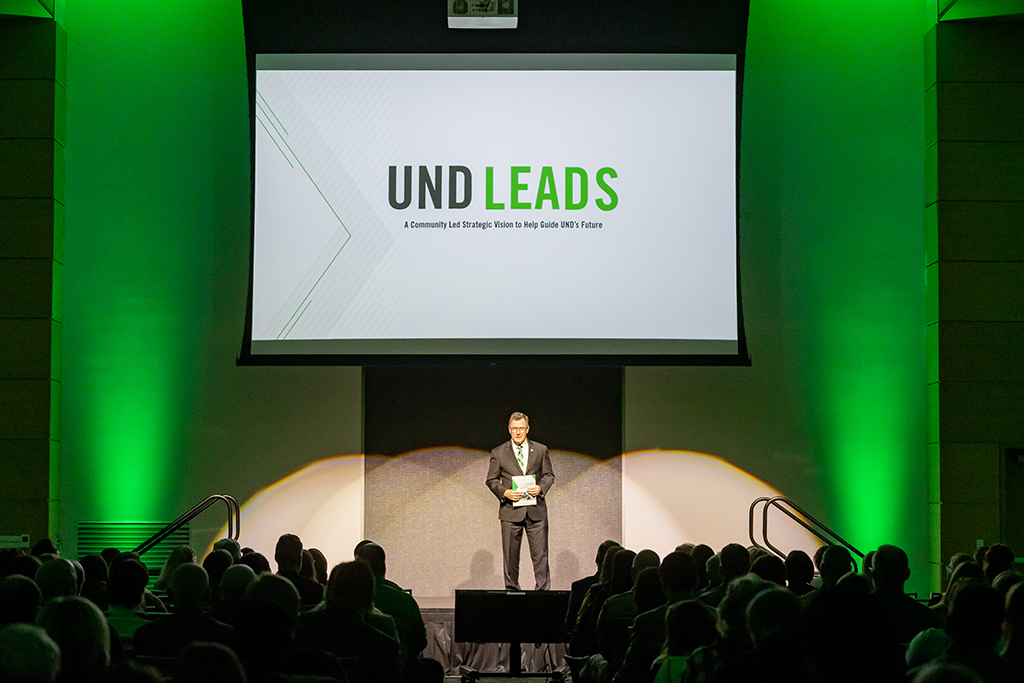 UND President Andy Armacost stands onstage beneath a large screen displaying the UND LEADS logo, with an audience seated in front of him.