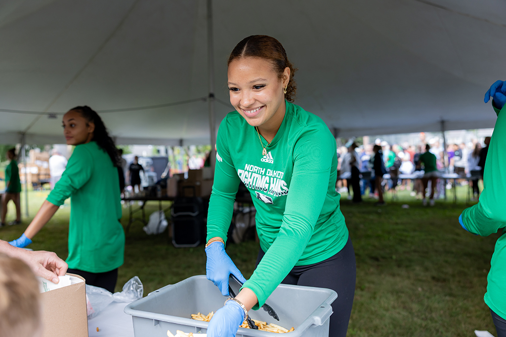 A smiling UND volunteer wearing a green Fighting Hawks shirt and blue gloves serves french fries from a large container.