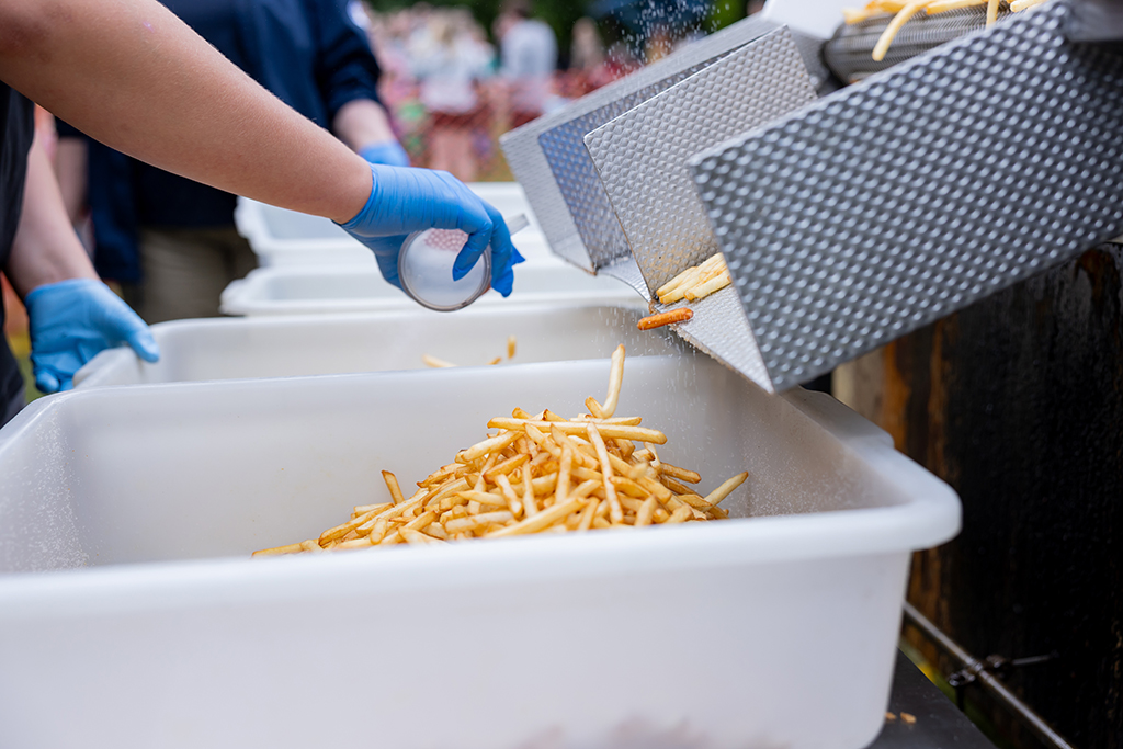 A volunteer wearing blue gloves pours freshly cooked french fries from a metal basket into a large container.
