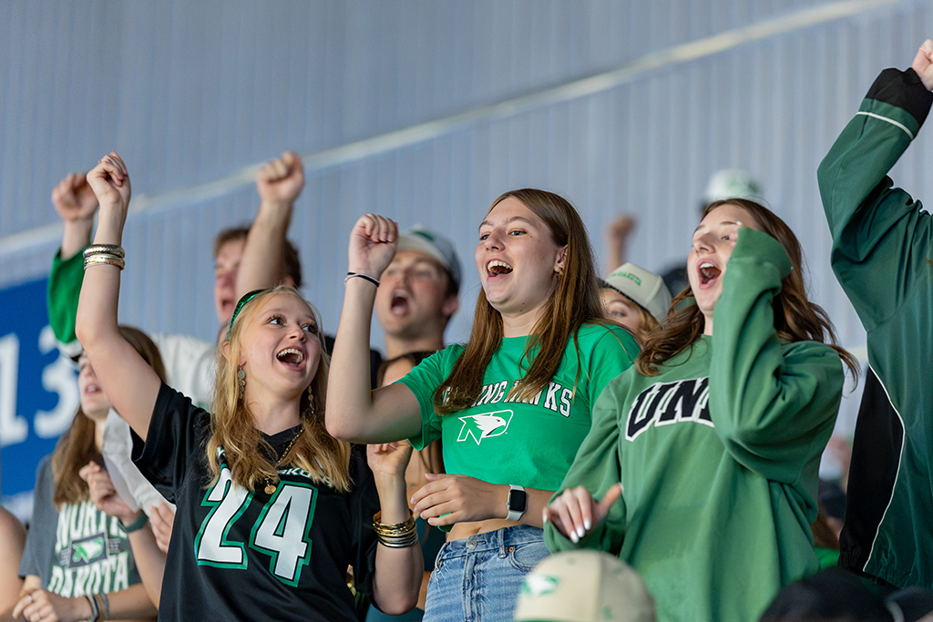 A group of students dressed in UND green and black cheer with their arms raised in the stands.