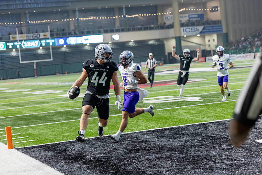 A UND football player carries the ball into the end zone with a St. Thomas defender pursuing him.