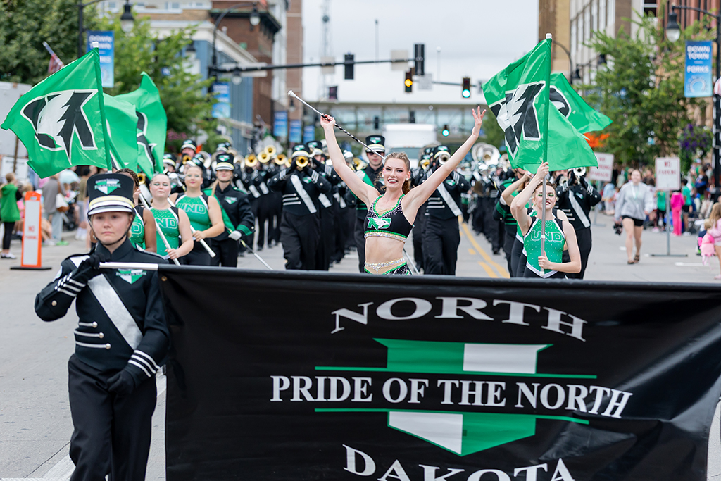 UND color guard members carrying green Fighting Hawks flags lead the Pride of the North marching band down a downtown street.