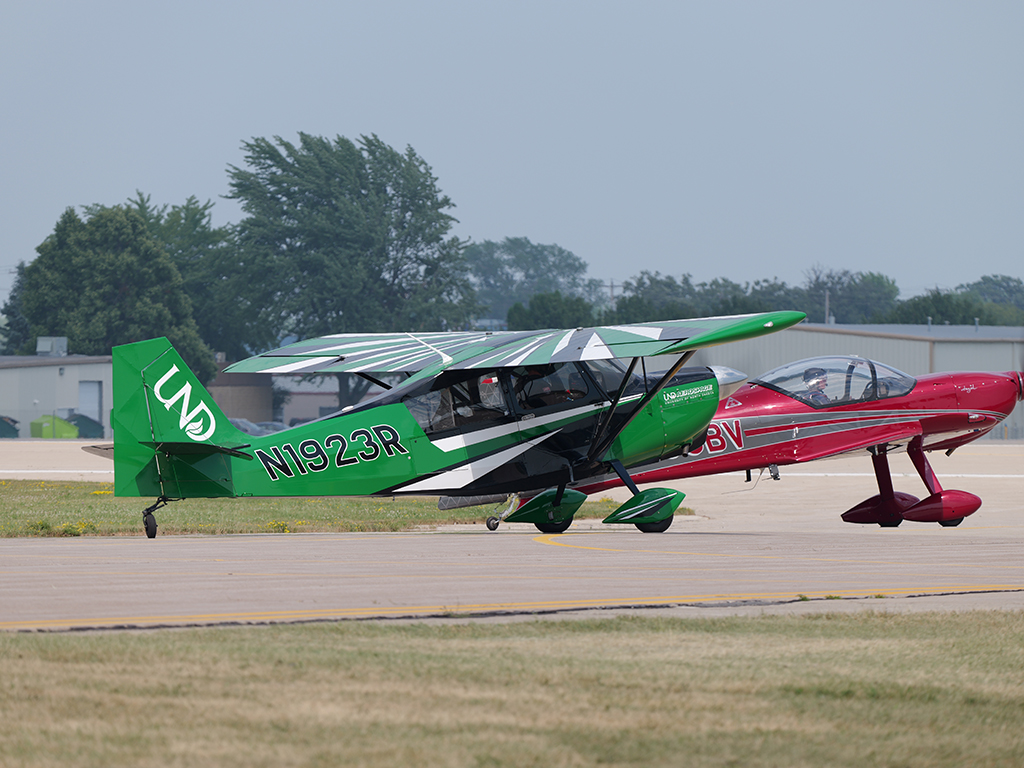 A green UND Aerospace American Champion Super Decathlon and a red aerobatic aircraft sit side by side on a taxiway at EAA AirVenture Oshkosh.