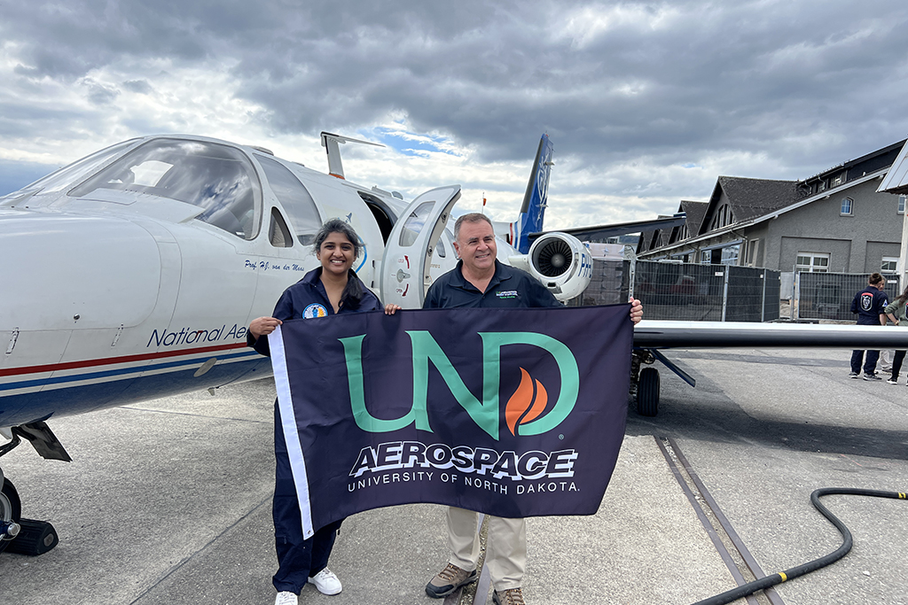 Pranika Gupta and Pablo de Leon hold a UND Aerospace flag in front of the research aircraft used for the parabolic flight campaign.