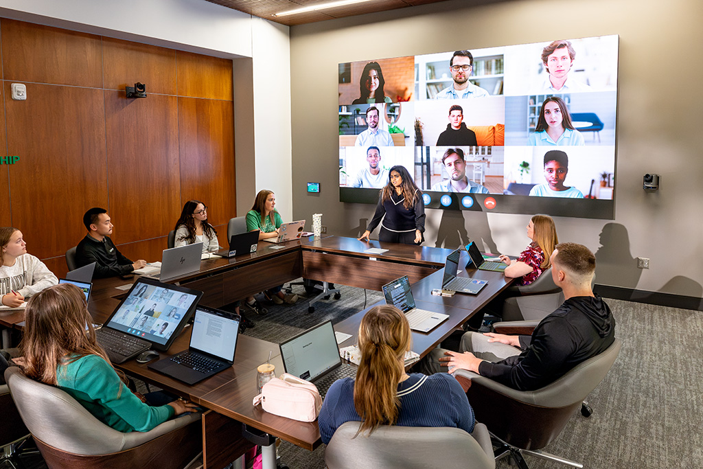 Students seated around conference tables with laptops participate in a hybrid learning session as a woman stands at the front of the room and remote participants appear on a large video screen.