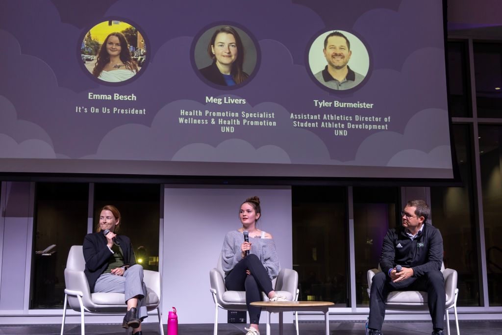 Three panelists sit in white chairs onstage holding microphones.