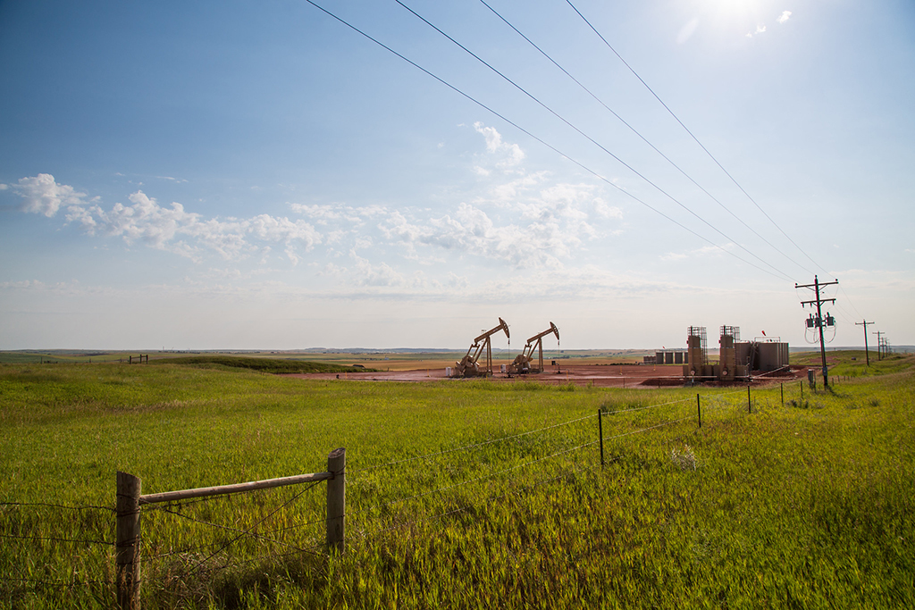 Two pumpjacks and oil production equipment sit in a grassy field beneath power lines in western North Dakota.