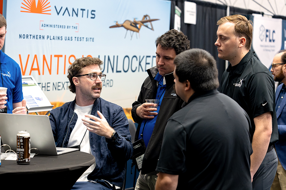 A seated exhibitor gestures beside an open laptop as several attendees gather around a table.