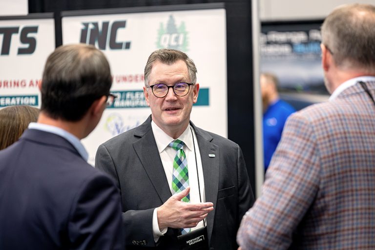 Andrew Armacost speaks with two attendees standing in front of exhibit booths.
