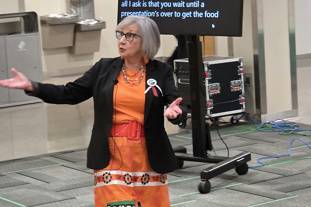 Dr. Ramona Klein gestures with both hands while speaking at UND's Indigenous Peoples' Day event. She wears a black blazer over an orange dress with ribbon skirt detailing and colorful beaded jewelry.