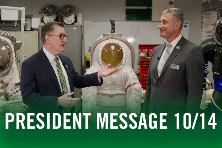 Andy Armacost and Bob Kraus in conversation near spacesuits with text in foreground: President Message 10/14